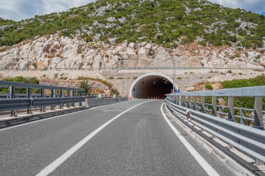 Her iki tarafında bariyerler olan iki şeritli bir yol bulutlu bir gökyüzünün altında yeşil bitkilerle kaplı büyük, kayalık bir tepeye oyulmuş bir tünele çıkıyor..