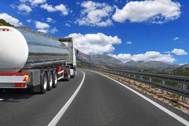 Parlak, gümüş renkli bir tanker, etrafı dağlarla çevrili ve beyaz bulutlu mavi gökyüzü ile çevrili, asfalt bir yolda ilerliyor..