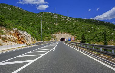 Her iki tarafında bariyerler olan iki şeritli bir yol bulutlu bir gökyüzünün altında yeşil bitkilerle kaplı büyük, kayalık bir tepeye oyulmuş bir tünele çıkıyor..
