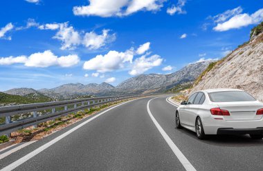 Beyaz bir araba, parlak mavi bir gökyüzünün altında kıvrımlı bir dağ yolu boyunca gidiyor. Dağılmış bulutlar manzaralı ve maceralı bir yolculuk atmosferi yaratıyor..