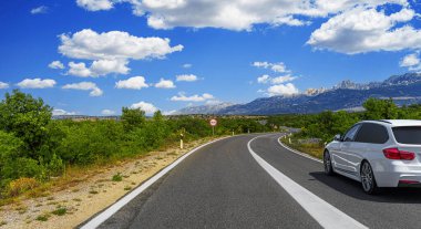 A white car drives along a winding mountain road surrounded by greenery and scenic views under a clear blue sky with clouds.