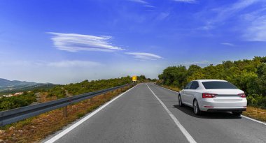 A white car drives along a winding mountain road surrounded by greenery and scenic views under a clear blue sky with clouds.