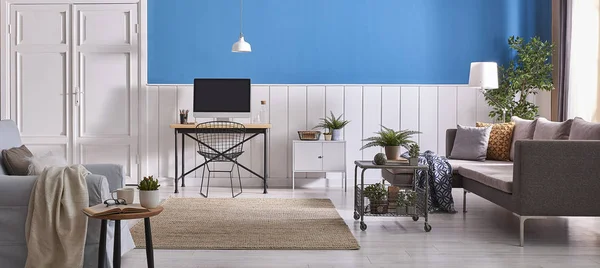 Modern living room, white and blue wall background, wooden working table ,grey sofa,computer and blue armchair . White lamp and green plants