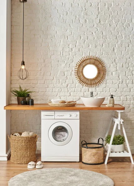 Washing machine in the laundry room, wooden table and shelf style, sink lamp mirror and wicker basket decor object.