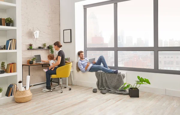 Anne baba ve bebek oturma odasında vakit geçiriyor, anne çalışıyor ve baba tablet bakıyor..