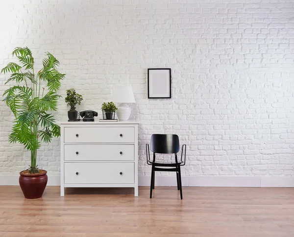 Decorative white cabinet and black chair in front of the brick wall, lamp, frame and vase of plant decorative style.