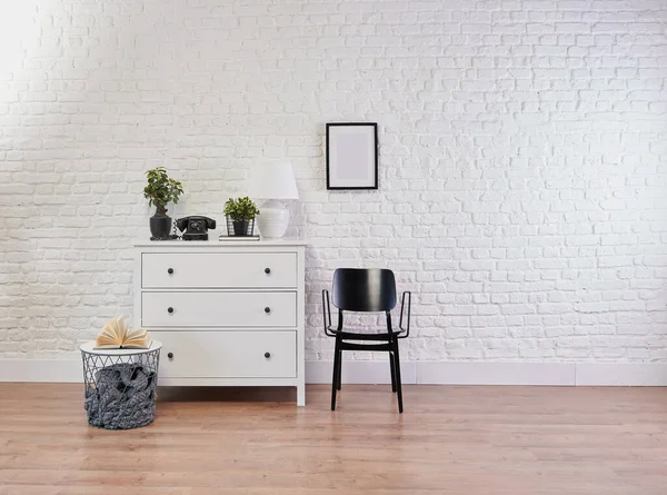 Decorative white cabinet and black chair in front of the brick wall, lamp, frame and vase of plant decorative style.