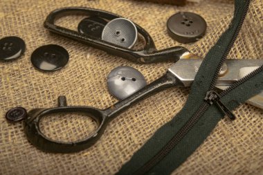 Çeşitli boyutlarda düğmeler, bir makara iplik, terzi makası ve kaba desenli çuval bezi üzerine bir fermuar. Kapat.