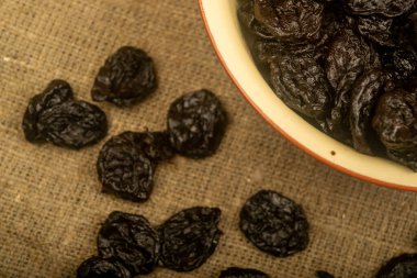 Seramik bir kasede kurutulmuş kuru erik ve kabarık desenli çuval bezi üzerine serpiştirilmiş kuru erik. Kapat.