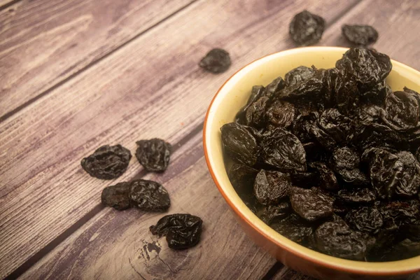 Dried prunes in a ceramic bowl and dried prunes scattered on a wooden background. Close up