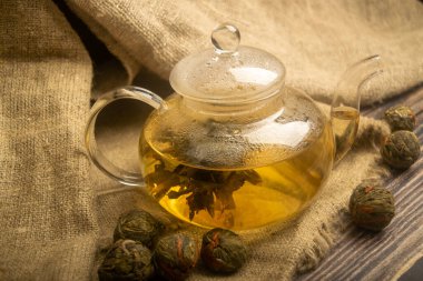 Cam çaydanlıkta demlenmiş bir çiçek çayı. Kabataslak desenli ev yapımı kumaştan bir arka planda. Kapat.