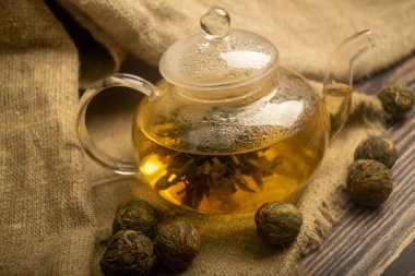 Cam çaydanlıkta demlenmiş bir çiçek çayı. Kabataslak desenli ev yapımı kumaştan bir arka planda. Kapat.