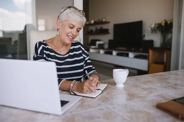 Mature beautiful woman making notes while working on her laptop