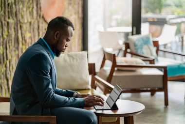 Modern bir ofisin lobisindeki masada oturan ve dijital tablet üzerinde çalışan Afrikalı genç bir işadamı.