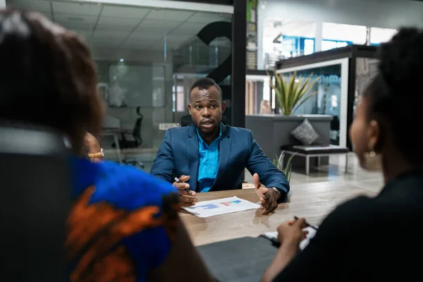 オフィスの役員室のテーブルの周りで一緒に会議中に同僚のグループと話している若いアフリカのビジネスマン — ストック写真