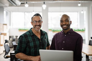 İki gülümseyen iş adamının portresi. Parlak, modern bir ofiste yan yana duruyorlar ve bir dizüstü bilgisayar üzerinde çalışıyorlar.