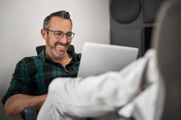 Smiling mature businessman sitting with his feet up on his desk and working online with a laptop