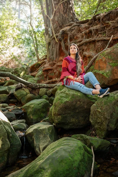 Woman deep in thought while sitting alone on some moss covered rocks by a small stream in a forest