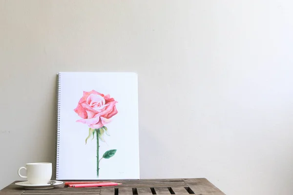 Pink rose by color pencil drawing on white paper. Hand drawing flower illustration on wooden table leaning on beige wall with color pencil and white coffee cup