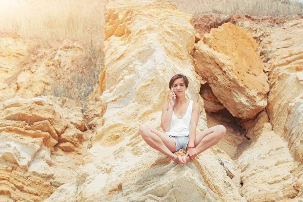 Una hermosa joven con el pelo corto está vestida con pantalones cortos y un jersey blanco está practicando yoga en el fondo de las rocas. Pose of the lotus. El concepto de calma y concentración — Foto de Stock