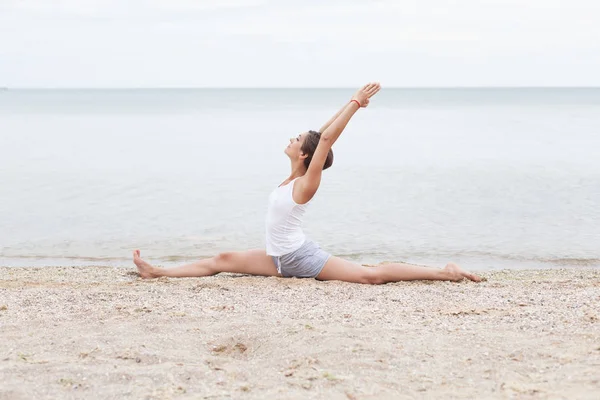 A girl with a short hairstyle dressed in shorts and a white jersey makes a twine on the background of the sea and the beach. Playing sports in nature. Stretching, Yoga pose.