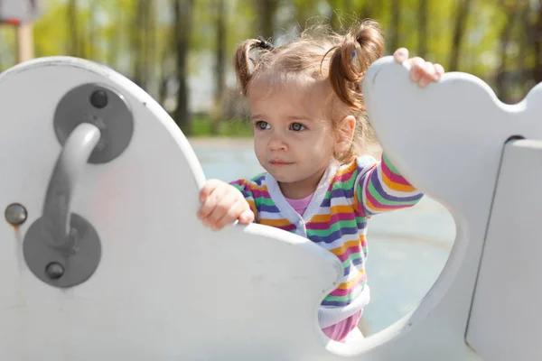 Een klein meisje met twee staarten is gekleed in een gestreepte kleurrijk jasje is spelen op de speelplaats — Stockfoto