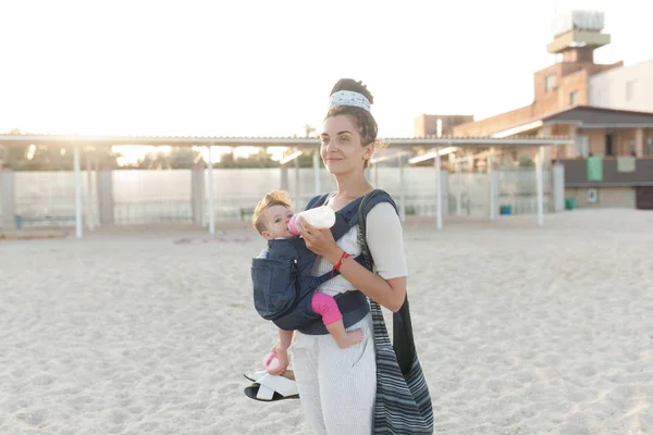 A small child sits in a backpack and walks along with the mother along the seashore. Summer family vacation concept — Stock Photo, Image