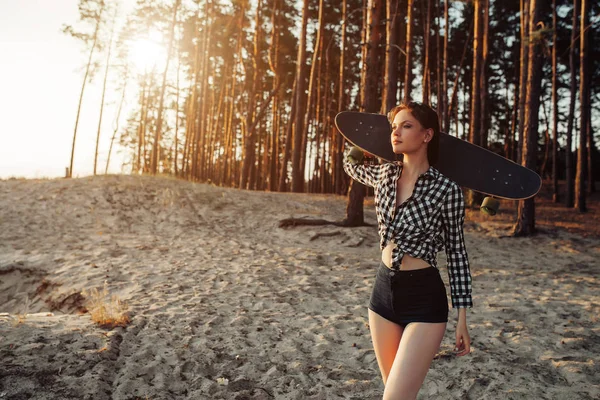 Una hermosa chica con un longboard en sus manos en la naturaleza en un bosque de pinos en busca de un buen camino que cabalgaría. Concepto de juventud estilo de vida moderno — Foto de Stock