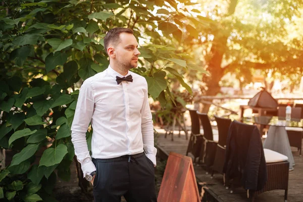Красивый молодой бизнесмен в белой рубашке и галстук бабочка на бизнес-ужин ждет своих коллег в одиночестве за летним столом ресторана — стоковое фото