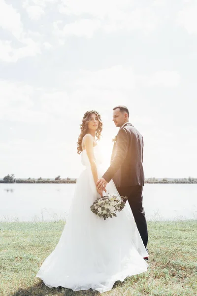 Casal em traje de casamento com um buquê de flores e vegetação está nas mãos contra o pano de fundo do campo ao pôr do sol, a noiva eo noivo — Fotografia de Stock