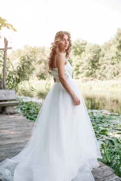 A cute curly woman in a white wedding dress with a wedding bouquet and wreath in her hair standing back to the camera in nature. Concept escaped bride. Forward to a happy bright future Runaway — Stock Photo, Image