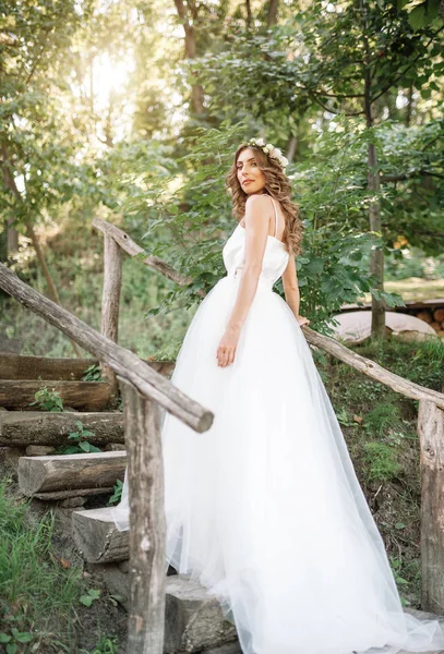 Eine niedliche, lockige Frau in einem weißen Brautkleid mit einem Brautstrauß und einem Kranz im Haar, die in der Natur in die Kamera blickt. Konzept entging Braut. Vorwärts in eine glückliche, strahlende Zukunft — Stockfoto