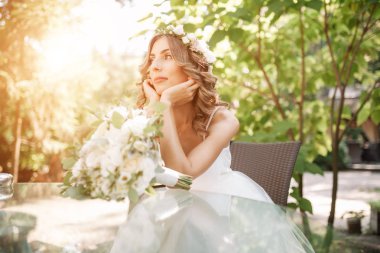 Genç gelin çene katlanmış elleriyle bir cam masaya oturur. Sıkılmış