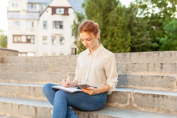 Belle jeune fille rousse avec des taches de rousseur assis sur un escalier et prendre des notes dans un carnet. Portrait d'un étudiant. Enseignement à distance. Travail à domicile — Photo