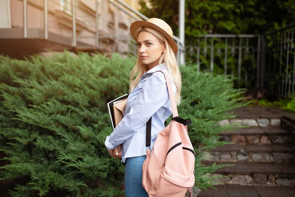Une belle étudiante blonde dans un chapeau de paille avec un sac à dos rose derrière son dos et des livres dans ses mains se tient devant l'entrée de l'université avec le dos à la caméra et souriant — Photo