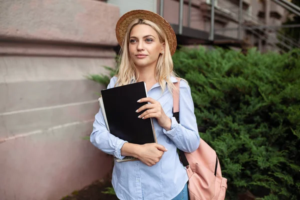 Счастливая красивая девушка улыбается и держит в руках стопку книг, стоящих рядом с кампусом. — стоковое фото