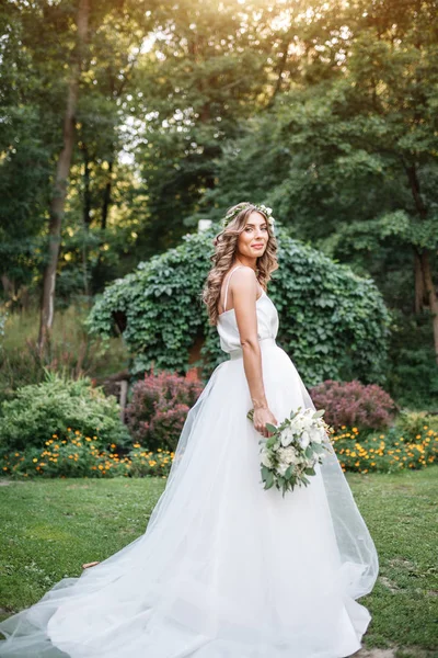 Eine niedliche, lockige Frau in einem weißen Brautkleid mit einem Brautstrauß und einem Kranz im Haar, die in der Natur in die Kamera blickt. Konzept entging Braut. Vorwärts in eine glückliche, strahlende Zukunft — Stockfoto