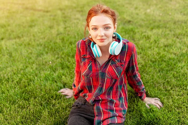 A beautiful red-haired student with freckles is dressed in a red checkered shirt with headphones sitting on the lawn in between the cheba. Student leisure — Stock Photo, Image
