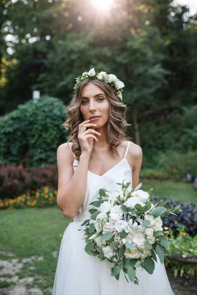 Schöne blonde glückliche Braut in elegantem weißen Kleid in einem Kranz mit Strauß im Freien — Stockfoto