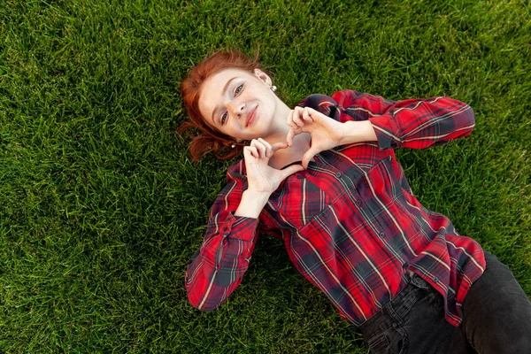 Beautiful red-haired young student with freckles lying on her back on the grass top view. Dressed in a red checked shirt, reveals a heart of fingers Love and positive — Stock Photo, Image