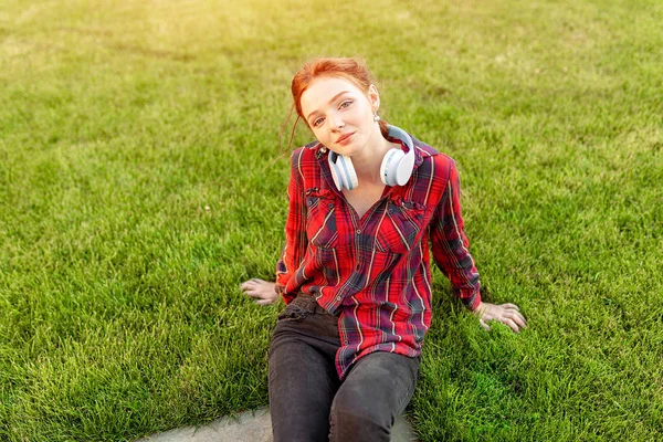 A beautiful red-haired student with freckles is dressed in a red checkered shirt with headphones sitting on the lawn in between the cheba. Student leisure — Stock Photo, Image