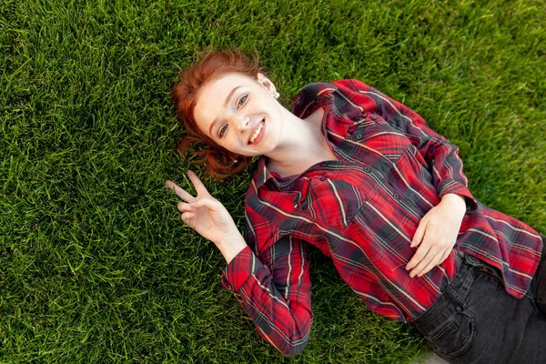 Beautiful young red-haired student girl with freckles lying on her back on grass and lawn view from above. Dressed in a red checkered shirt, resting after classes — Stock Photo, Image