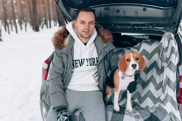 Jeune beau mec dans une forêt enneigée d'hiver se trouve dans le coffre de sa voiture dans une étreinte avec son ami chien de race Beagle — Photo