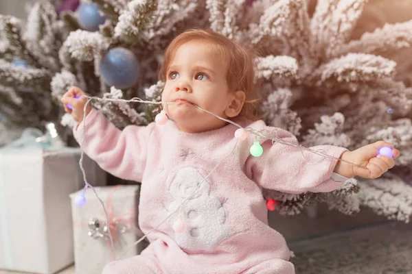 Small girl in the pink pajama sitting near the christmas tree playing with the garland — Stock Photo, Image