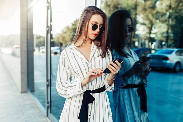 Счастливая привлекательная молодая женщина в солнечных очках смотрит на экран смартфона во время прогулки по городу. Одетые в стильную одежду . — стоковое фото