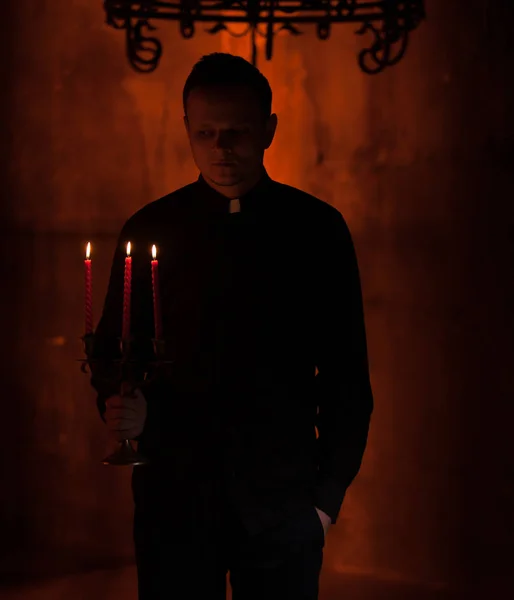 Joven sacerdote orante católico. Retrato del sacerdote Junto a las velas reza con las manos dobladas cerca de la cara. Pared roja en el fondo. Retrato guapo interior de la religión hombre — Foto de Stock