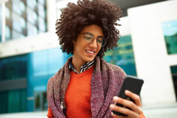 A handsome curly-haired guy of mixed race is dressed in a trendy youth-style scarf and sweater writes a message in a teleone against the background of office buildings.