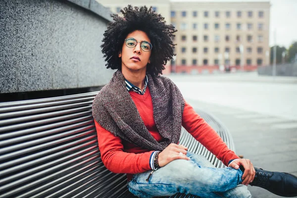 벤치에 앉아 잘생긴 젊은 이집트 곱슬 남자는 밝은 주황색 스웨터와 청바지 입고. 학생 수업 사이 휴식 시간에 산책. 귀여운 hipster 남자 — 스톡 사진