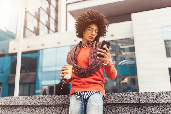 A handsome curly-haired guy of mixed race is dressed in a trendy youth-style scarf and sweater writes a message in a teleone against the background of office buildings.