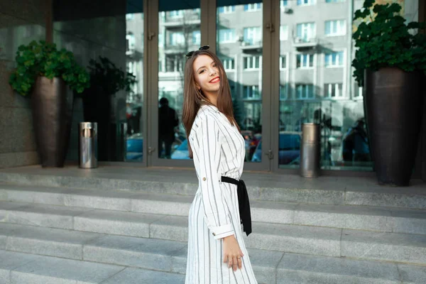 Jovem bonita alegre entrando porta de vidro no escritório moderno, hotel, café, centro de negócios e olhando para trás para acenar adeus. Menina vestindo óculos elegantes, e sorrindo de volta — Fotografia de Stock